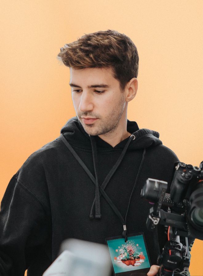 Young man with short brown hair wearing a black hoodie, looking down while holding a camera rig with an orange background.