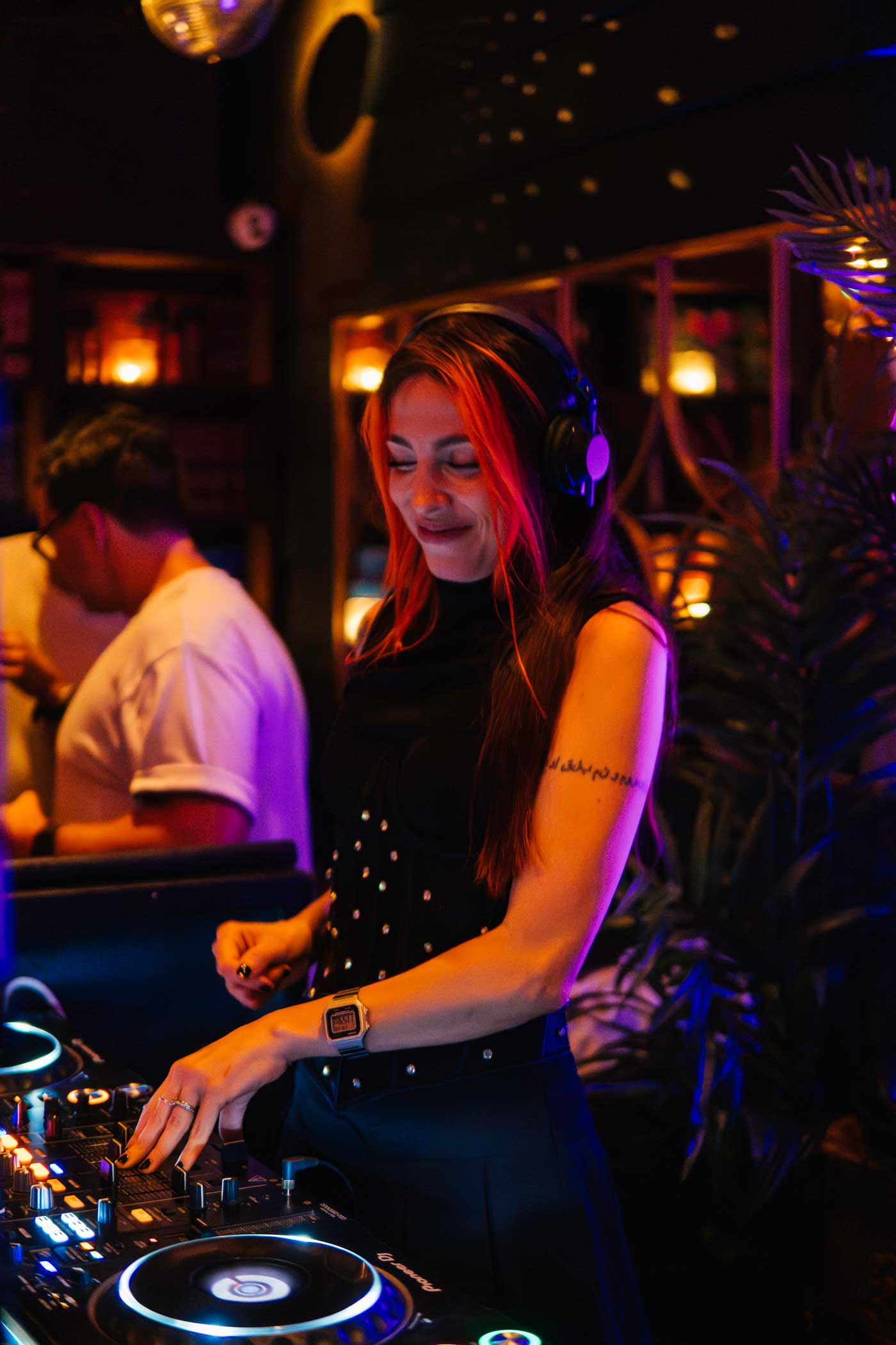 Woman with red hair and headphones DJing on a mixer in a dimly lit club with warm ambient lights.