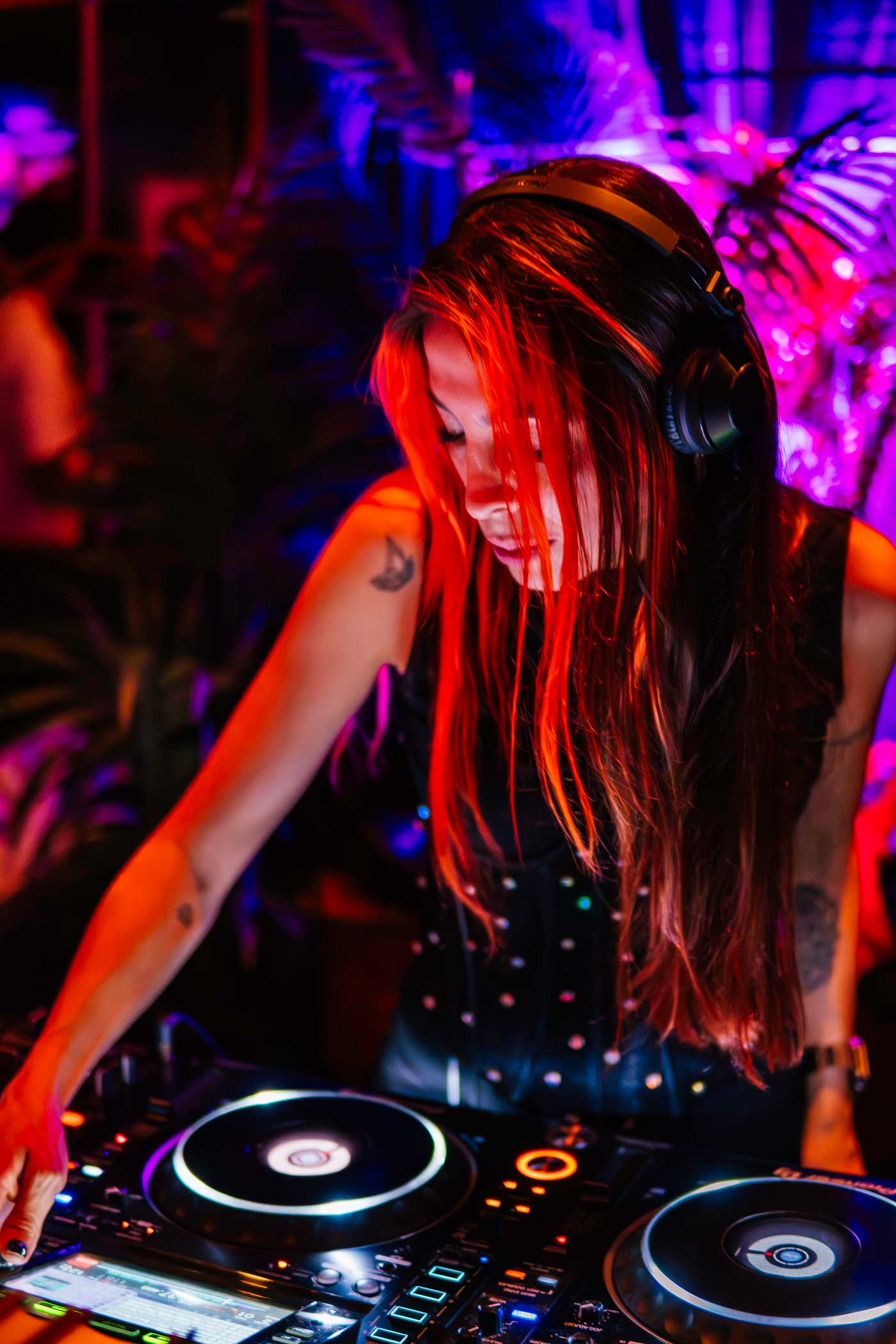 Female DJ with long red hair and headphones playing music on a DJ mixer in a colorful club setting.
